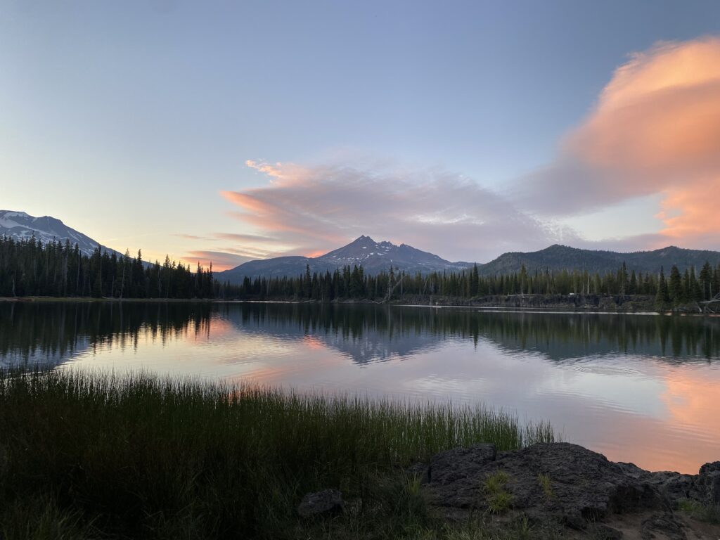 “Calm alpine lake reflecting mountain peaks and pine forest in Oregon at sunset