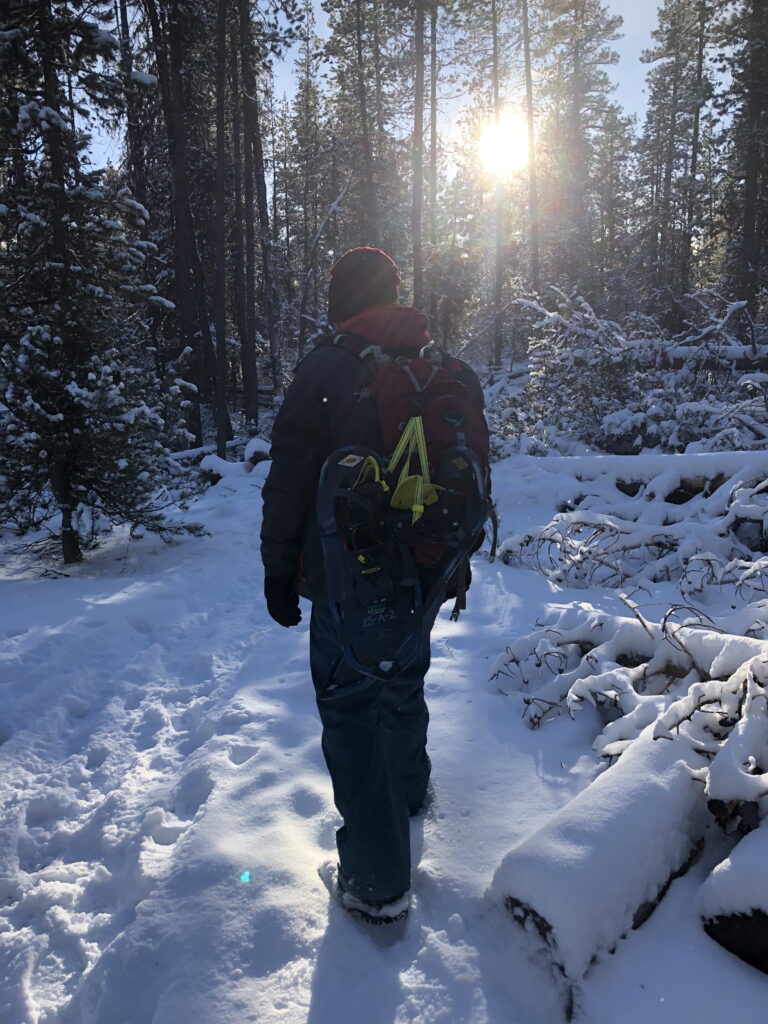 Hiker in Central Oregon carrying snowshoes on a backpack while walking along a snowy forest trail in winter.